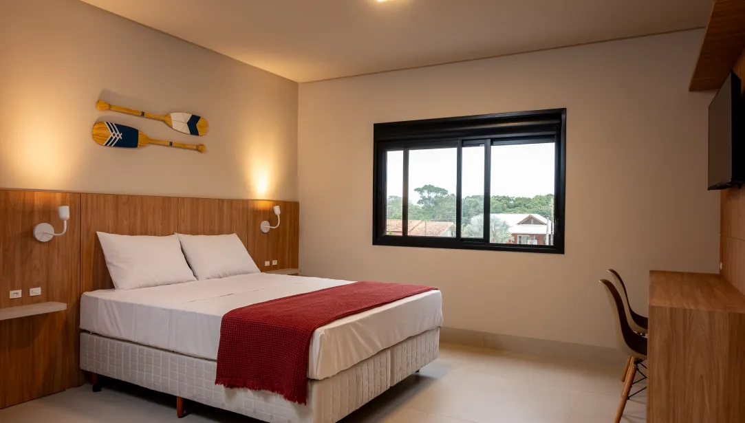 Quarto da pousada com cama de casal, decoração minimalista, manta vermelha e janela que oferece vista para a área externa da pousada.