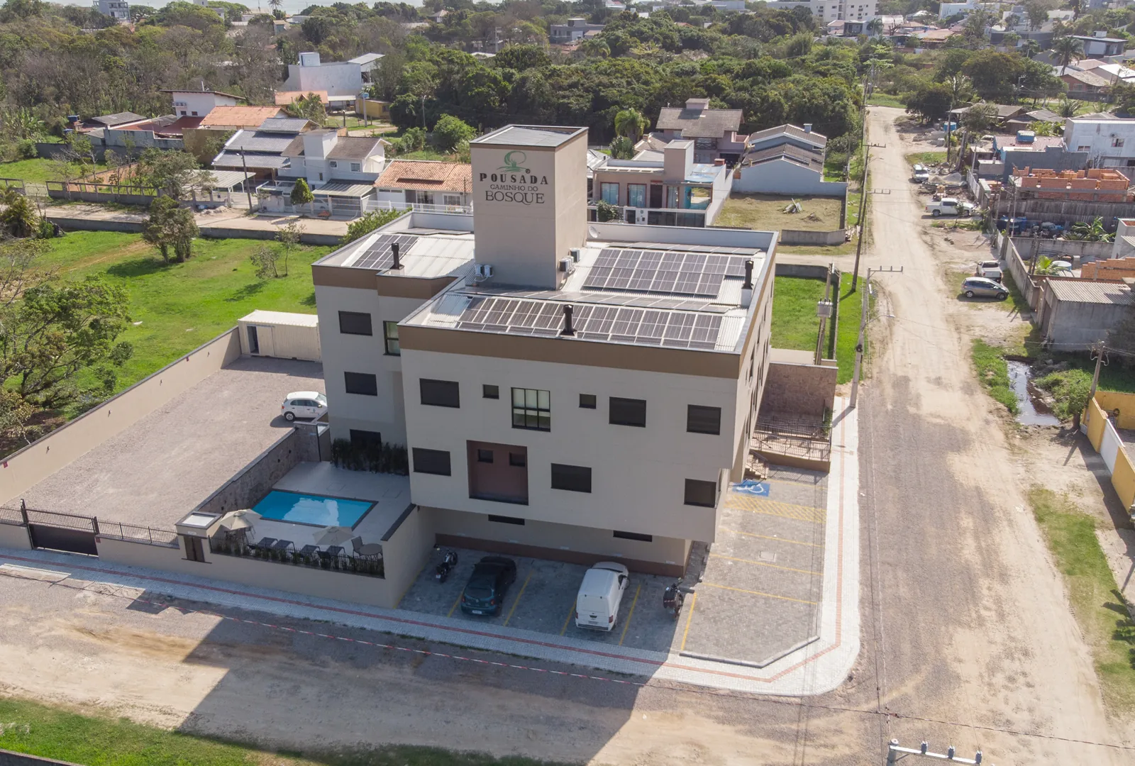 Vista aérea da Pousada Caminho do Bosque com estacionamento privativo, piscina e painéis solares no telhado, localizada em uma área tranquila de Bombinhas.