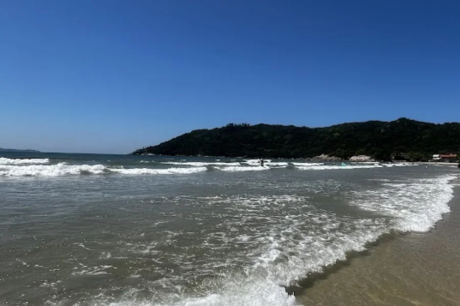 Vista da Praia de Mariscal em Bombinhas, com águas cristalinas, ondas moderadas e uma paisagem rodeada por montanhas verdes, ideal para relaxar e praticar esportes aquáticos.