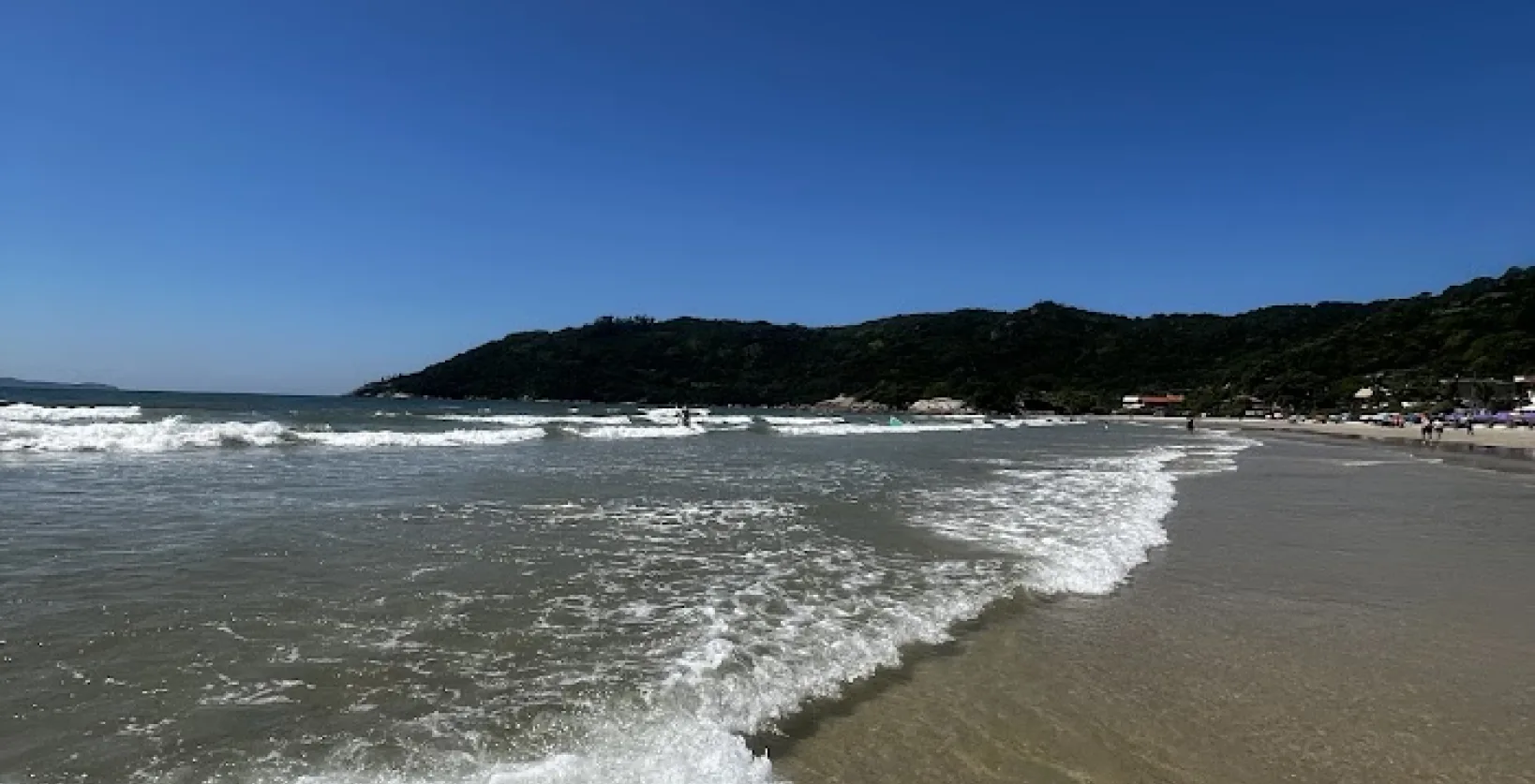 Vista da Praia de Mariscal em Bombinhas, com águas cristalinas, ondas moderadas e uma paisagem rodeada por montanhas verdes, ideal para relaxar e praticar esportes aquáticos.