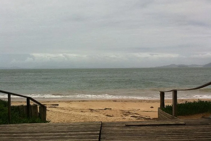 Vista da Praia da Tainha em Bombinhas, mostrando o acesso por um deck de madeira, areia dourada e mar calmo cercado por um cenário natural tranquilo.